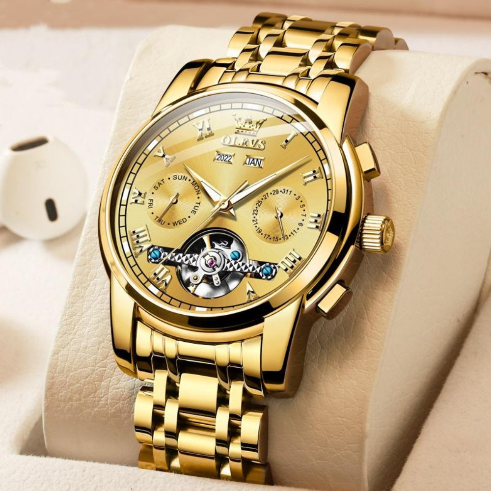 A men's luxury watch with a polished golden finish and Roman numerals, featuring a multifunction chronograph dial, exposed gear detail, and a stainless steel build.