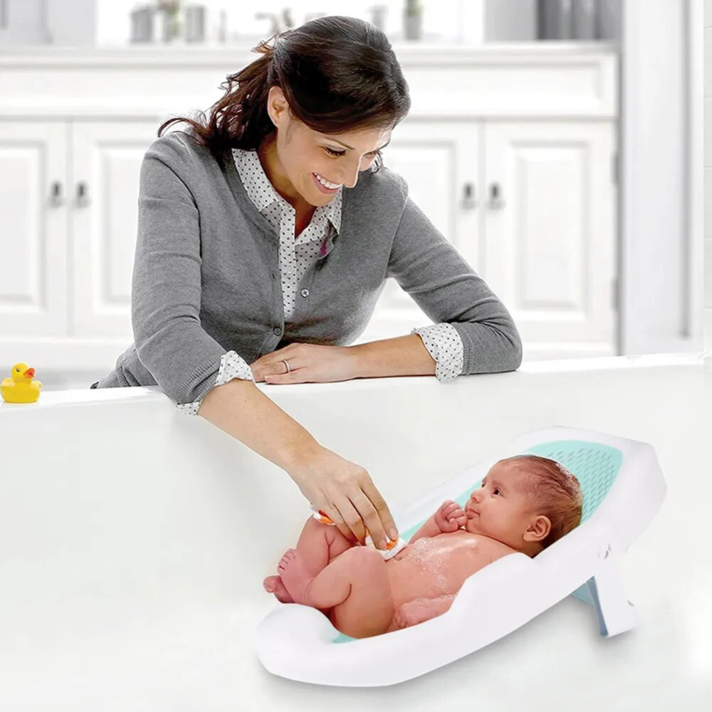 A baby is being bathed using the Anti-Slip Adjustable Baby Bath Support seat, which is white with a green trim. The baby is on the support seat, and an adult is smiling and reaching towards the baby with a duck toy.