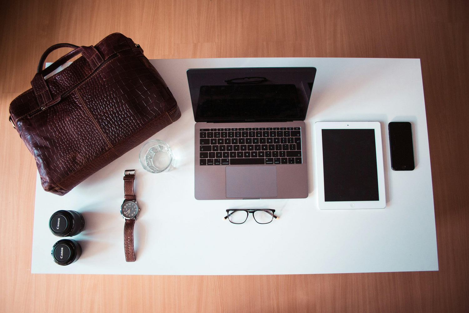 Flat lay of workspace with laptop, tablet, smartphone, glasses, brown leather bag, watch, camera lenses, and glass of water on white table