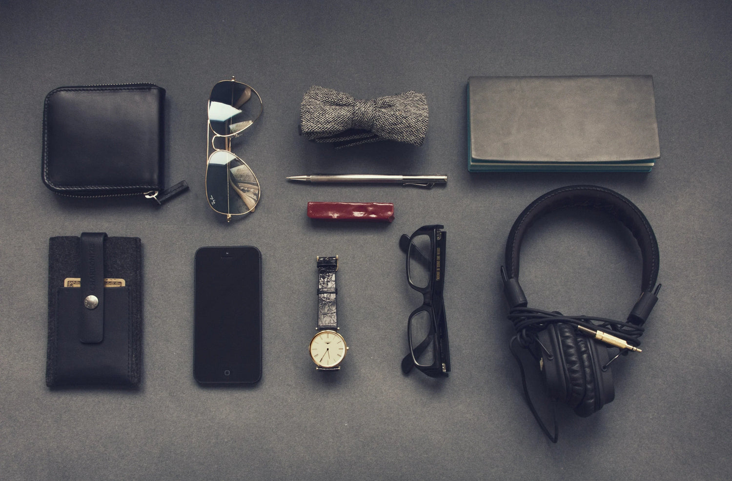 Flat lay of men's accessories including wallet, sunglasses, bow tie, pen, glasses, watch, smartphone, headphones on gray background