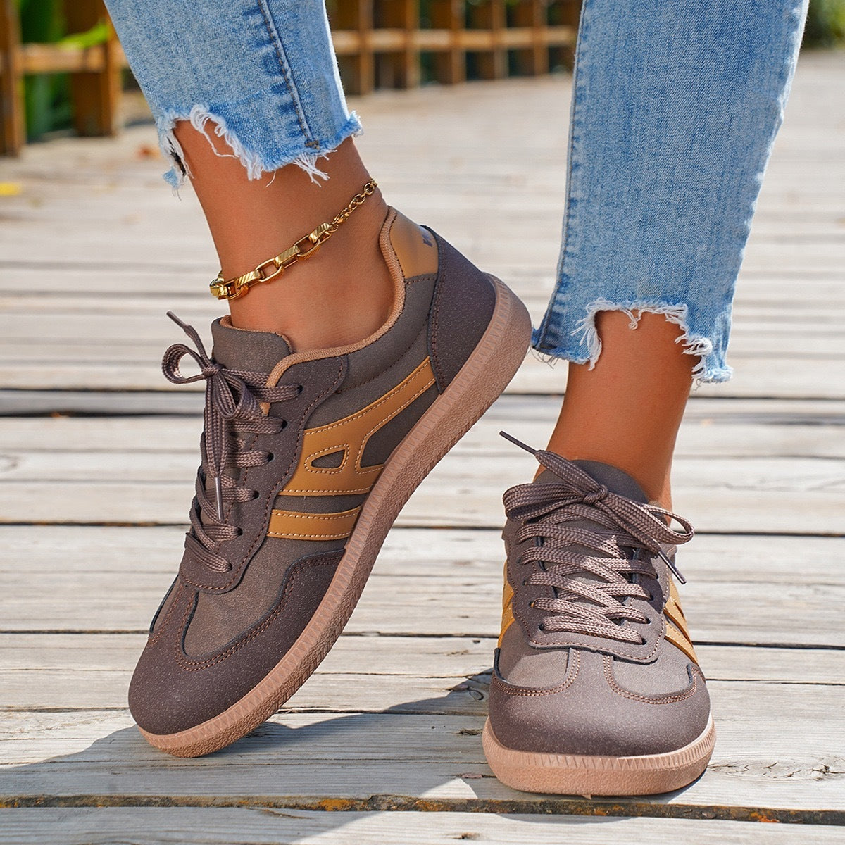 Close-up of brown and beige sneakers worn with ripped light blue jeans and a gold ankle bracelet on wooden deck