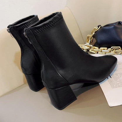 Black leather ankle boots with block heels on a beige surface next to a black bag with gold chain