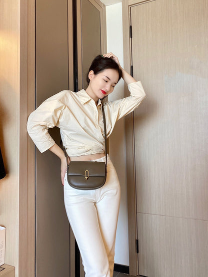 Woman in beige cropped shirt and white pants posing indoors with olive green shoulder bag