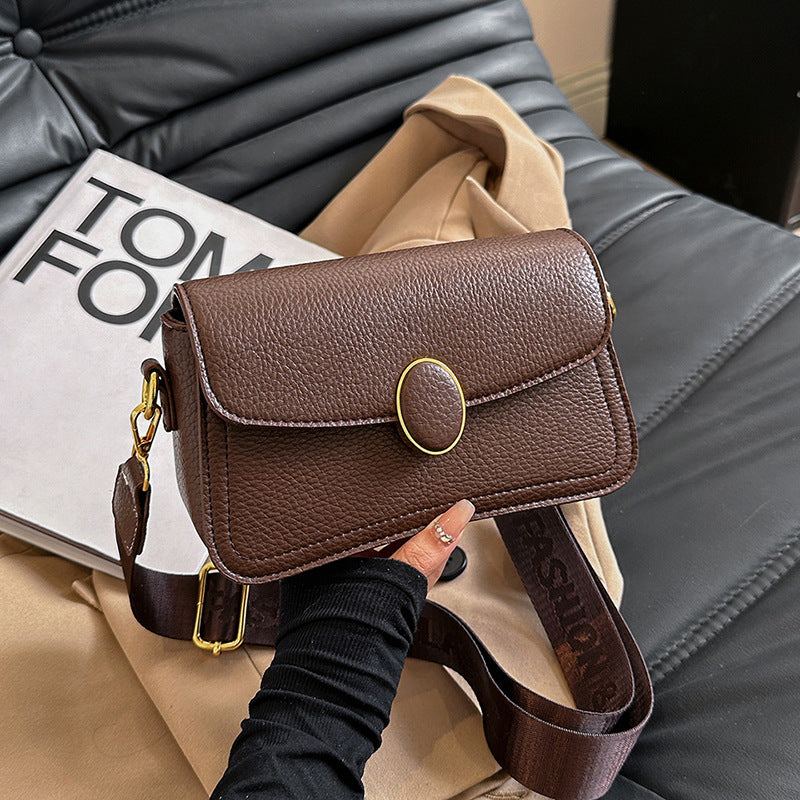 Brown textured leather crossbody bag with gold oval clasp and wide strap held over a tan and black background