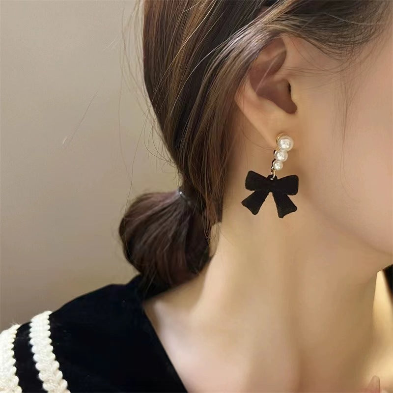 Black bow-shaped earring with pearl details on a person's ear against a neutral background