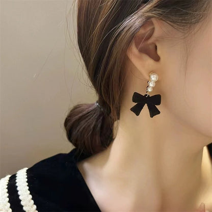 Black bow-shaped earring with pearl details on a person's ear against a neutral background