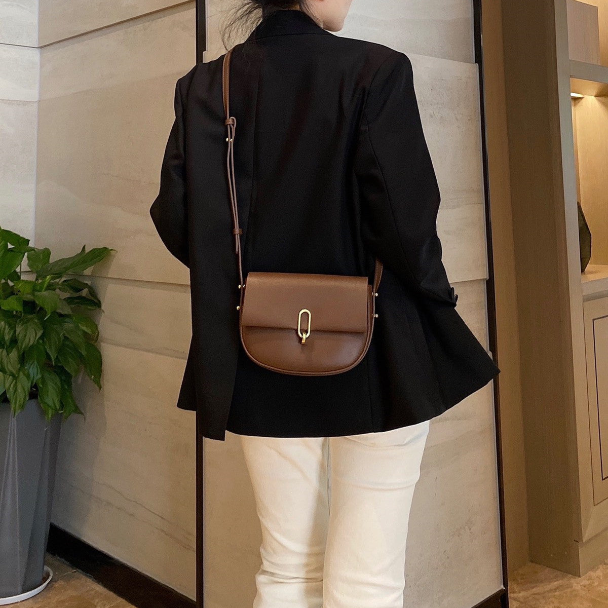 Woman in black blazer and white pants carrying a brown leather crossbody bag indoors