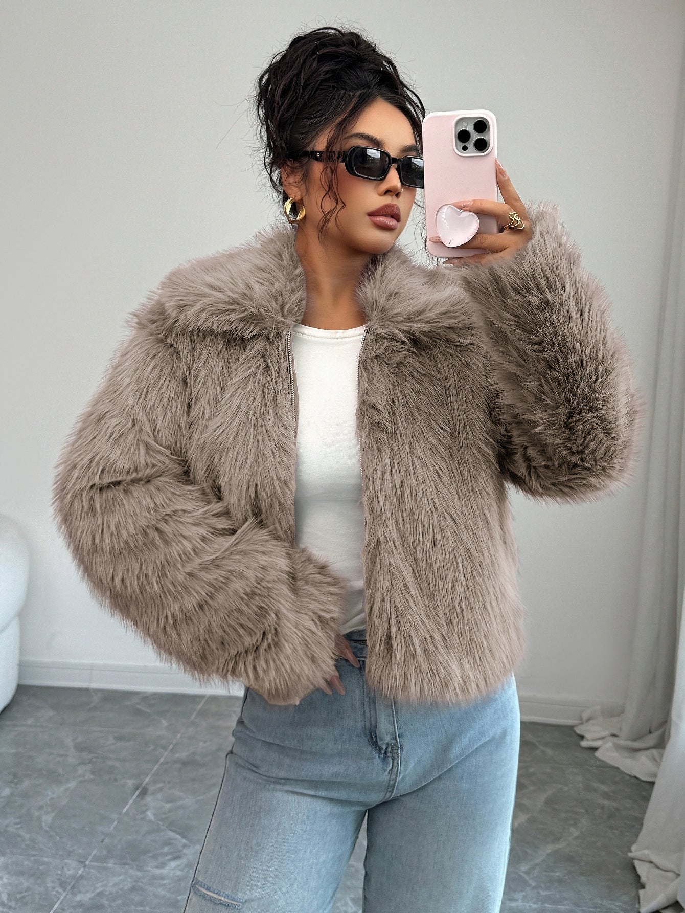 Woman wearing beige faux fur jacket, white top, blue jeans, black sunglasses, taking a selfie indoors