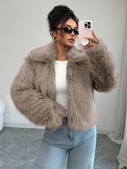 Woman wearing beige faux fur jacket, white top, blue jeans, black sunglasses, taking a selfie indoors