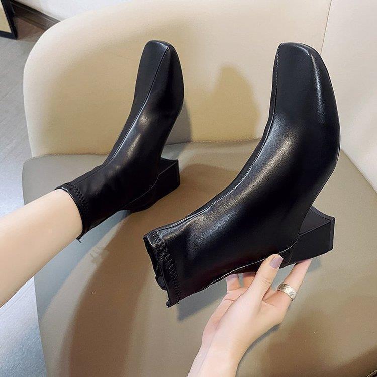 Pair of sleek black ankle boots with block heels on beige chair, one worn and one held