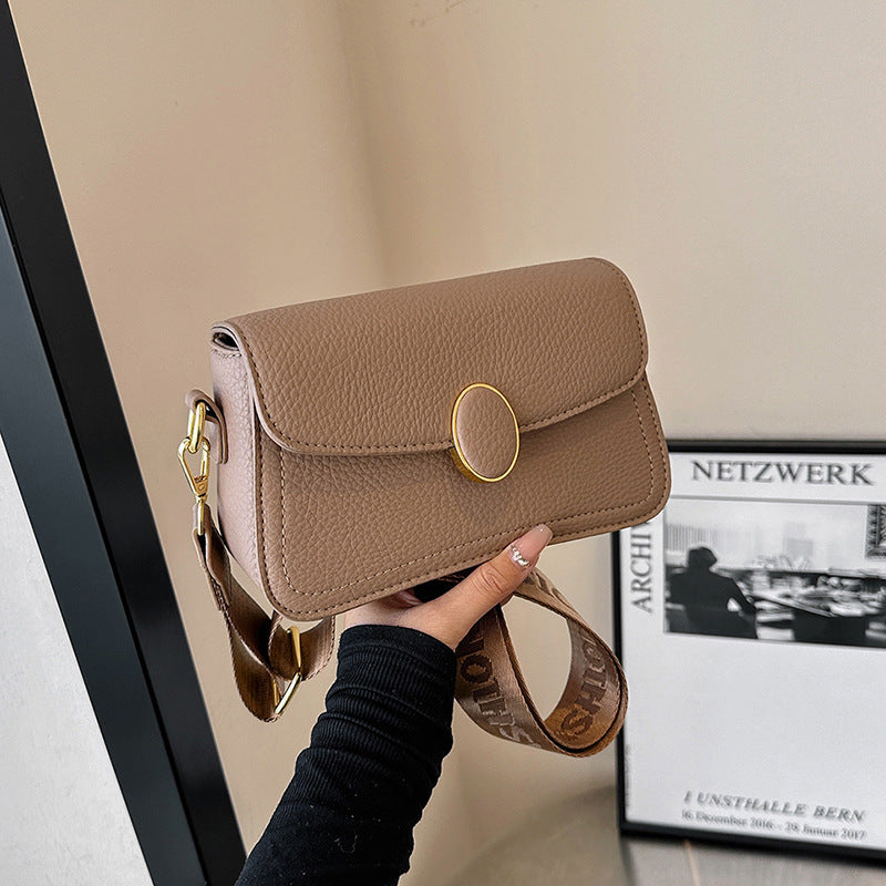 Hand holding taupe leather crossbody bag with gold oval clasp and adjustable strap in modern room