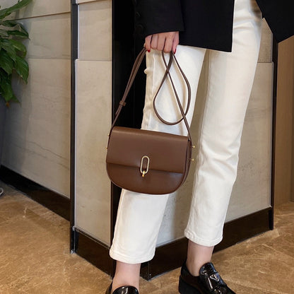 Woman holding brown leather crossbody bag wearing white pants and black shoes indoors