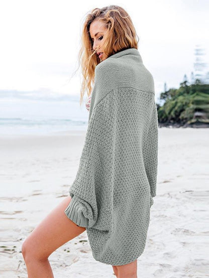 Woman wearing oversized gray knit cardigan standing on a sandy beach with ocean and greenery background