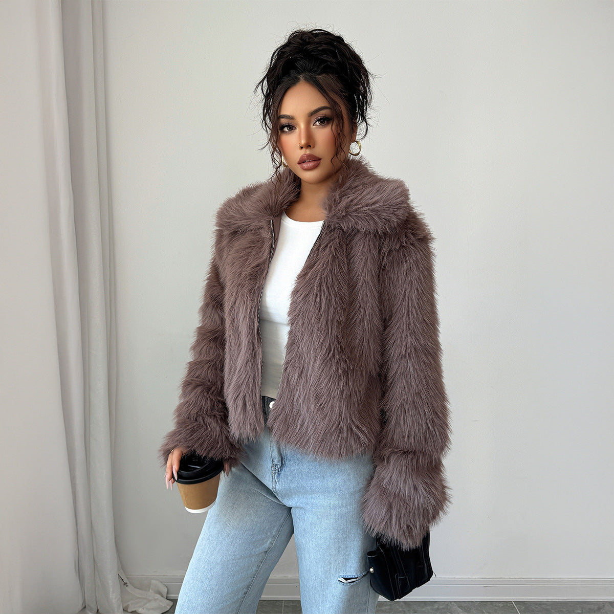 Woman in a mauve faux fur jacket, white top, and light blue jeans holding coffee cup and black purse