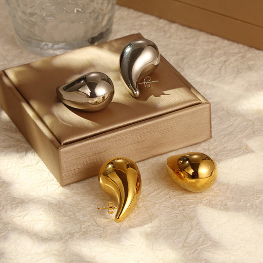 Shiny silver and gold teardrop-shaped stud earrings on a satin beige jewelry box with soft natural light