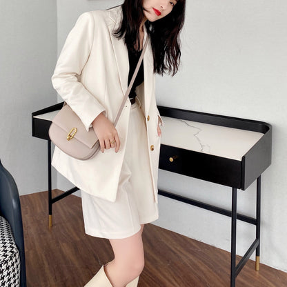Woman in cream blazer and shorts with beige purse, black top, and white boots indoors