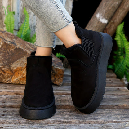 Black slip-on winter boots with fur lining worn with light blue jeans on wooden surface with greenery background