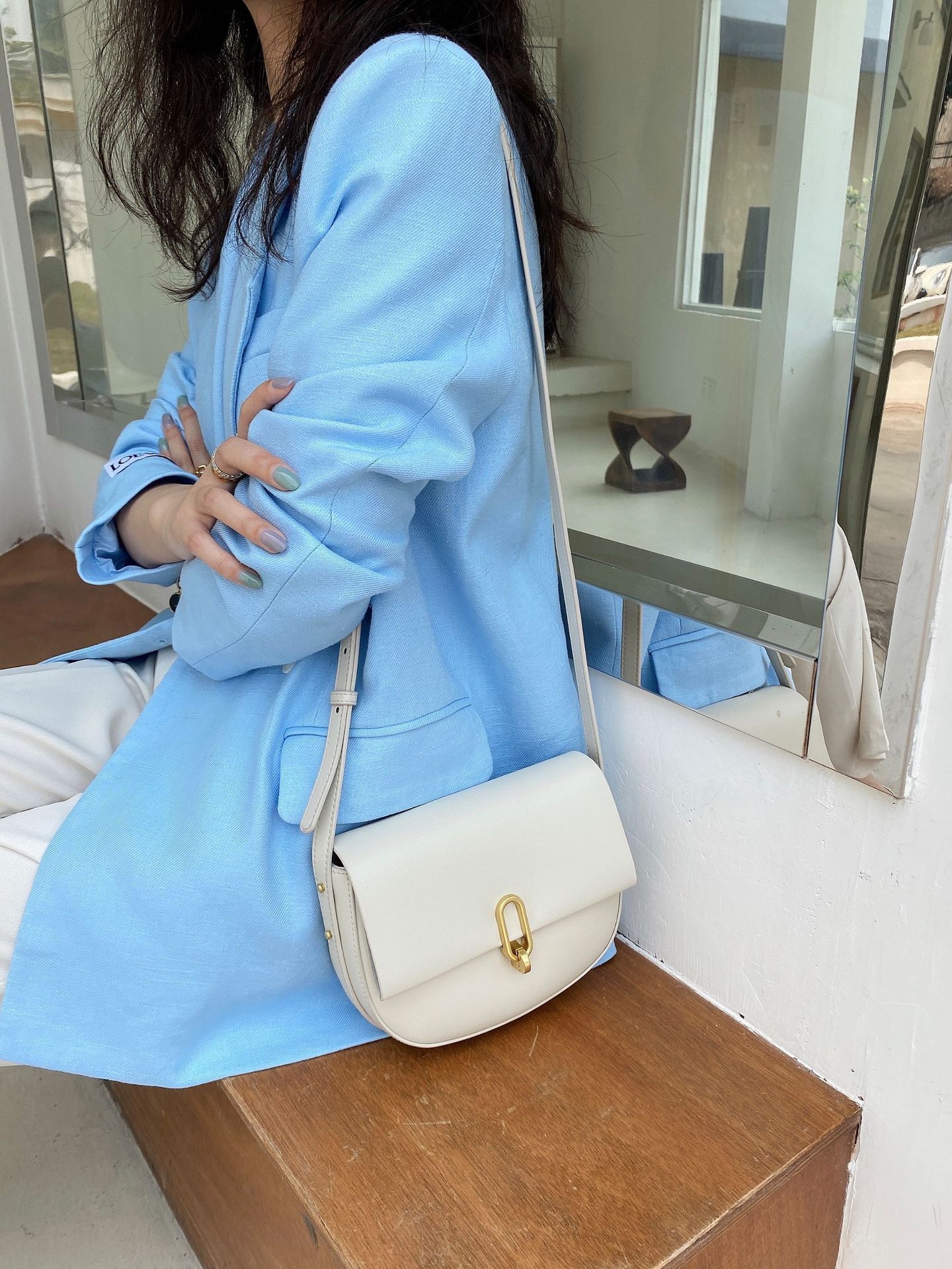 Woman wearing light blue blazer and white pants sitting on wooden bench with white leather shoulder bag
