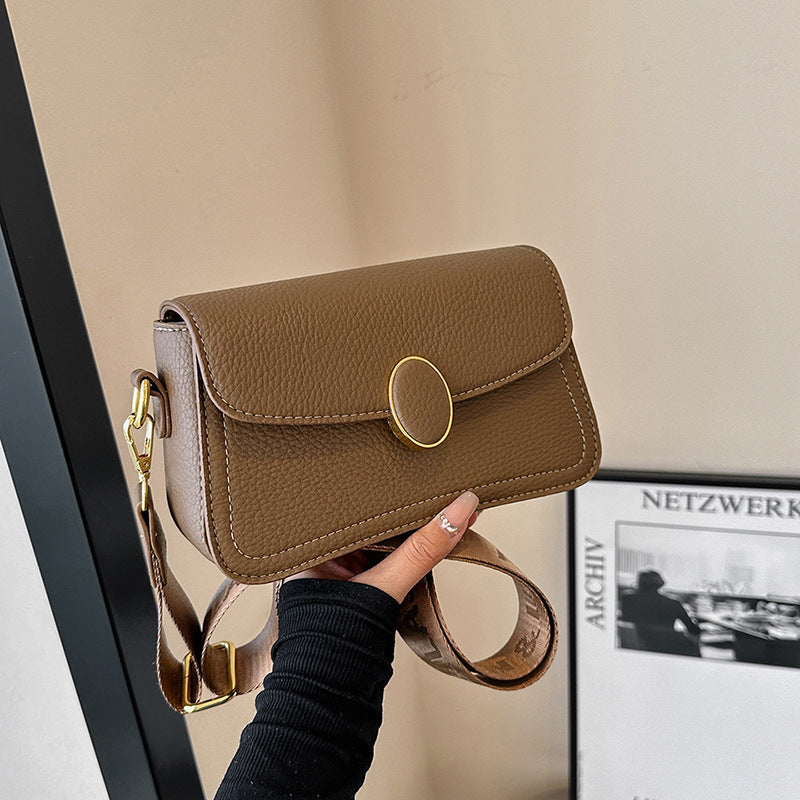 Hand holding a brown textured leather crossbody bag with a gold oval clasp and wide strap