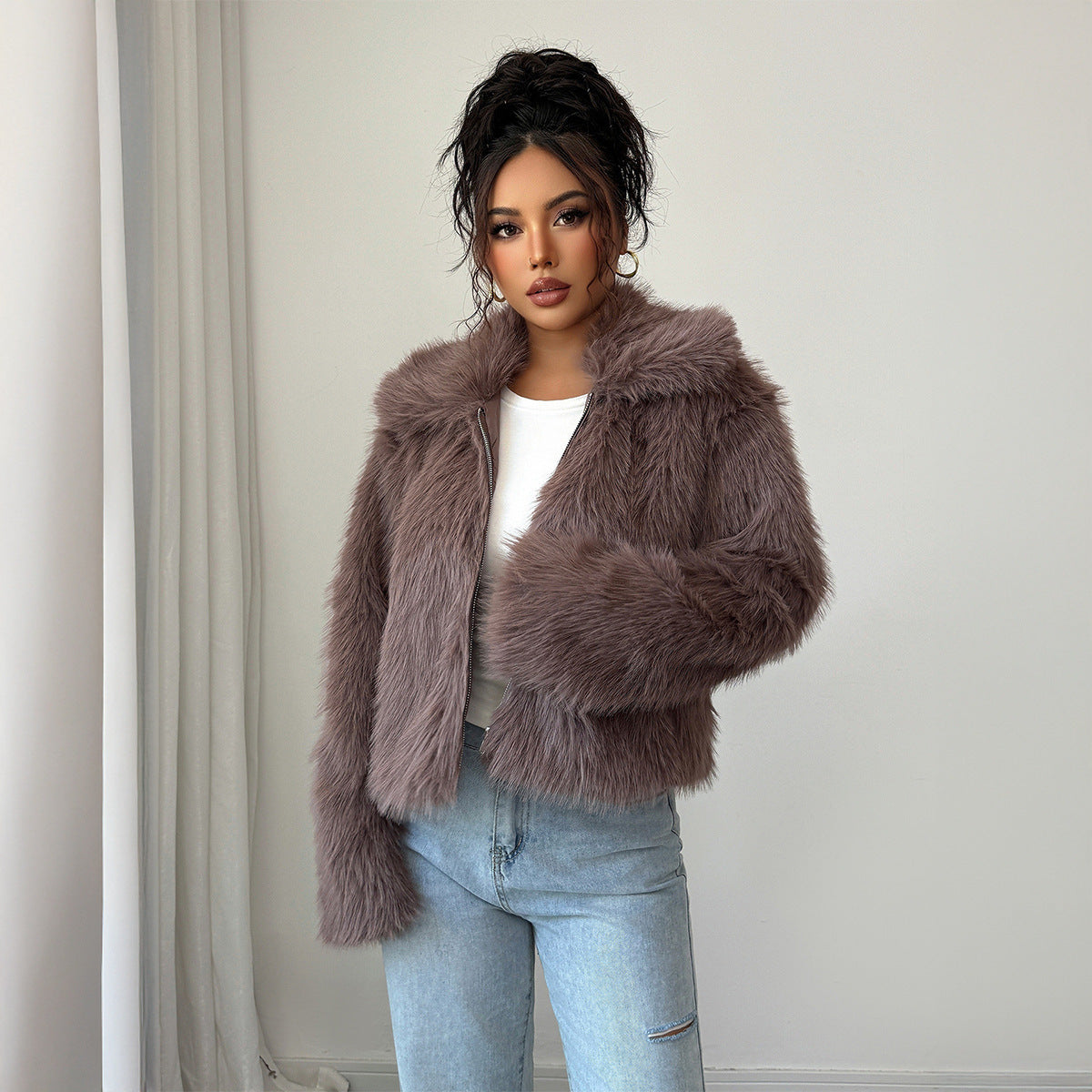 Woman with curly updo wearing mauve faux fur jacket over white top and light blue jeans indoors