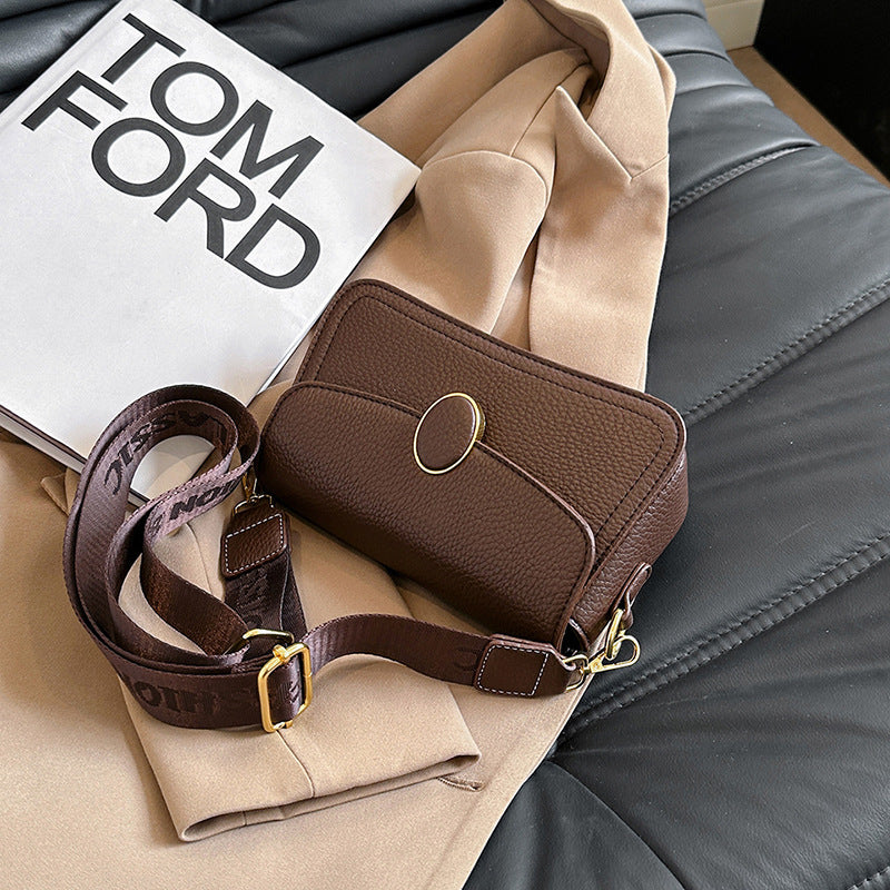 Brown leather crossbody bag with gold oval clasp on beige coat and Tom Ford book on black leather surface