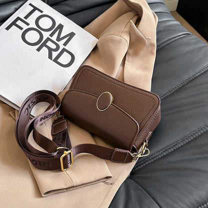 Brown leather crossbody bag with gold oval clasp on beige coat and Tom Ford book on black leather surface